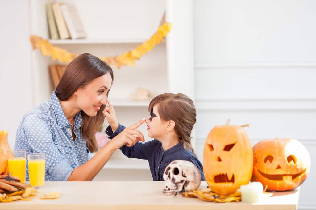 A mother painting her daughter's face at home in front of a table with Halloween decorations